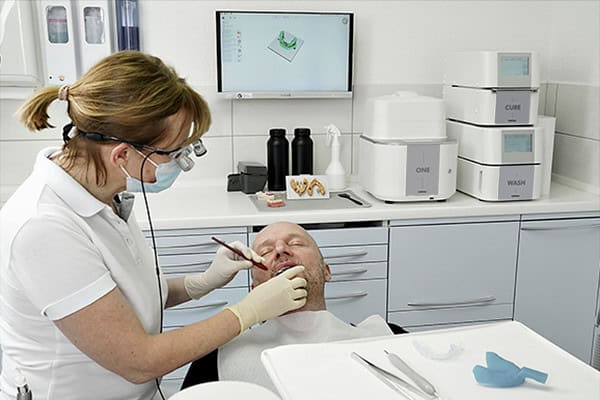 The ONE 3D Printing Solution, including the ONE wash and the ONE cure, RS Print Studio, and dental resins in a clinic, while the doctor works on a patient
